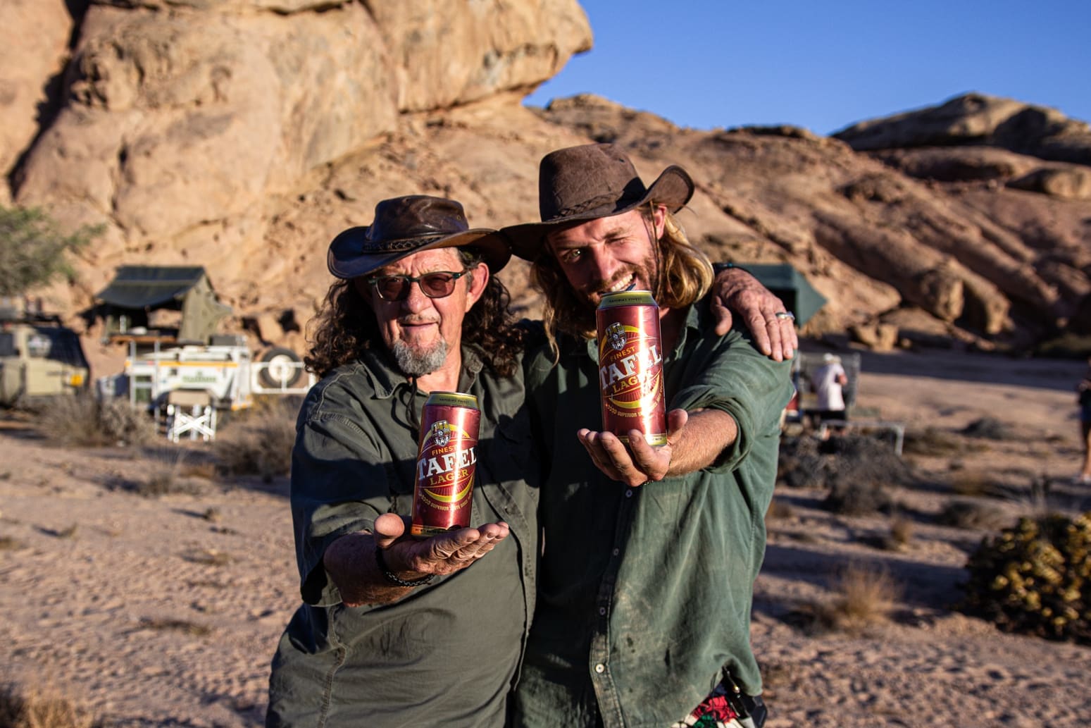 Twee mannen met hoeden en zonnebrillen staan buiten in een rotsachtig, woestijnachtig gebied, glimlachen en houden blikjes Tafel Lager bier in de richting van de camera. Op de achtergrond zijn terreinwagens en tenten te zien.