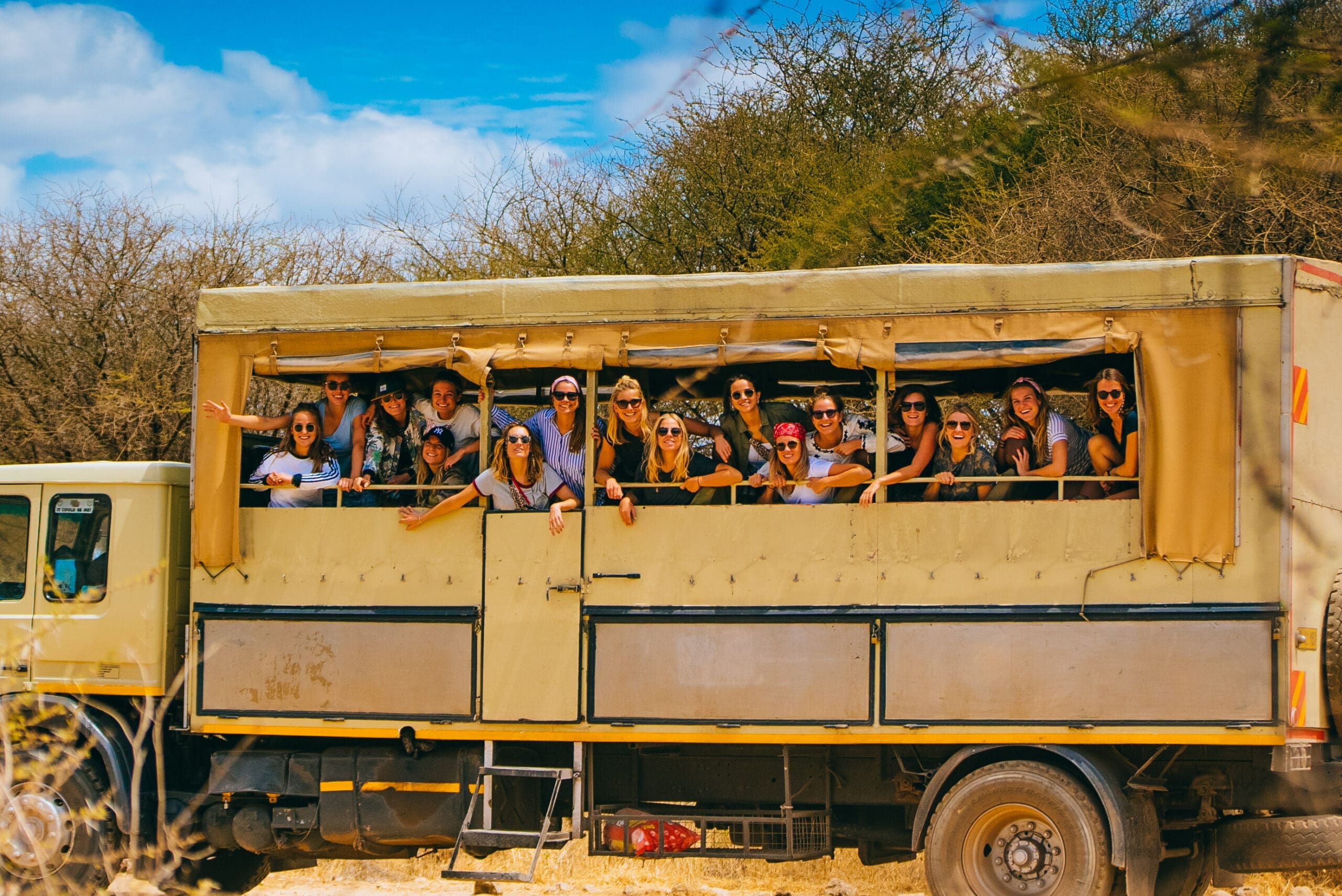 Een groep mensen glimlacht en leunt uit de ramen van een grote beige safaritruck die buiten geparkeerd staat, omringd door droge struiken onder een felblauwe hemel.