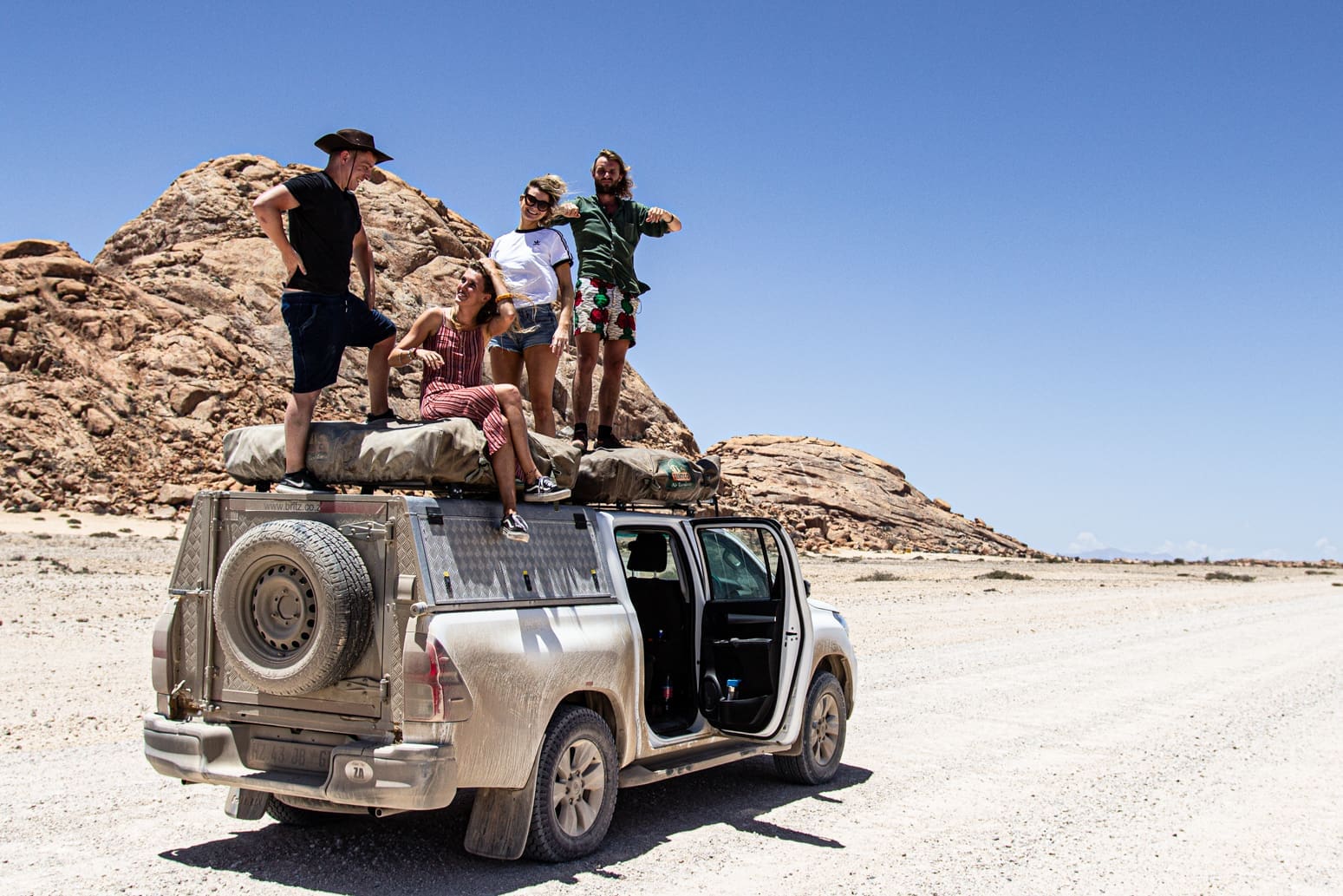 Vier mensen staan en zitten bovenop een stoffige SUV die geparkeerd staat op een zandweg in een rotsachtig woestijnlandschap onder een strakblauwe hemel. De autodeuren staan open en de groep lijkt te genieten van het landschap.