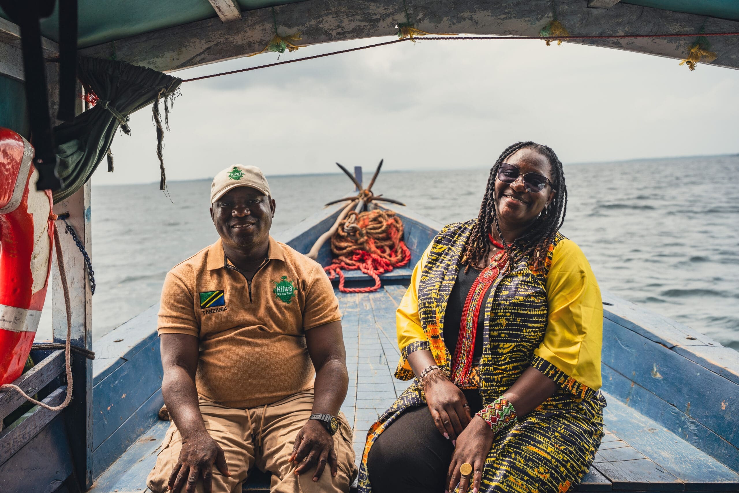 Twee mensen zitten glimlachend op een houten boot met opgerolde touwen achter zich, drijvend op kalm water onder een bewolkte hemel. De man draagt een bruin overhemd en een pet, terwijl de vrouw kleurrijke kleding met patronen en een zonnebril draagt.