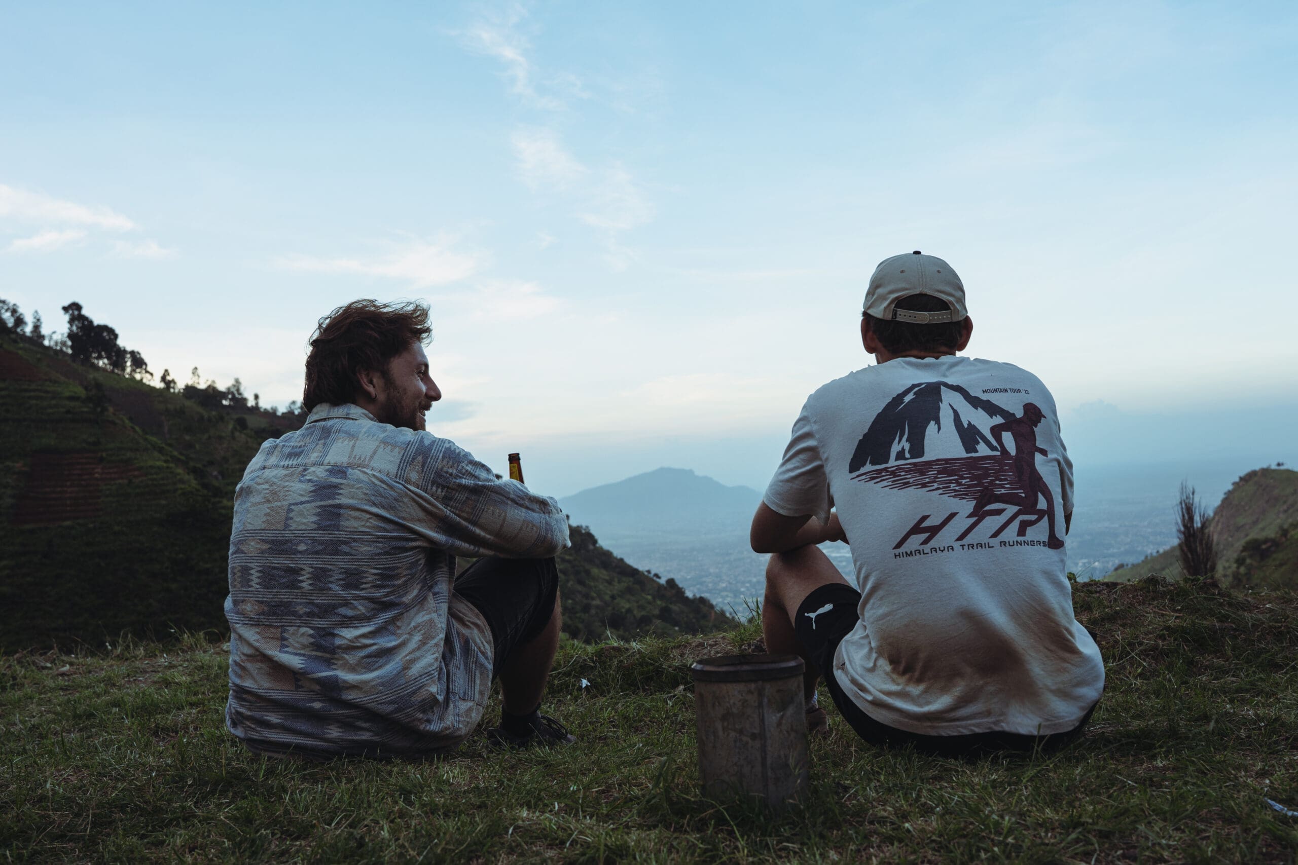 Twee mensen zitten op een grasheuvel met hun gezicht naar de andere kant, uitkijkend over een verre vallei en bergen in de schemering. Eén persoon houdt een fles vast en de ander draagt een pet en een shirt met een afbeelding van een berg.