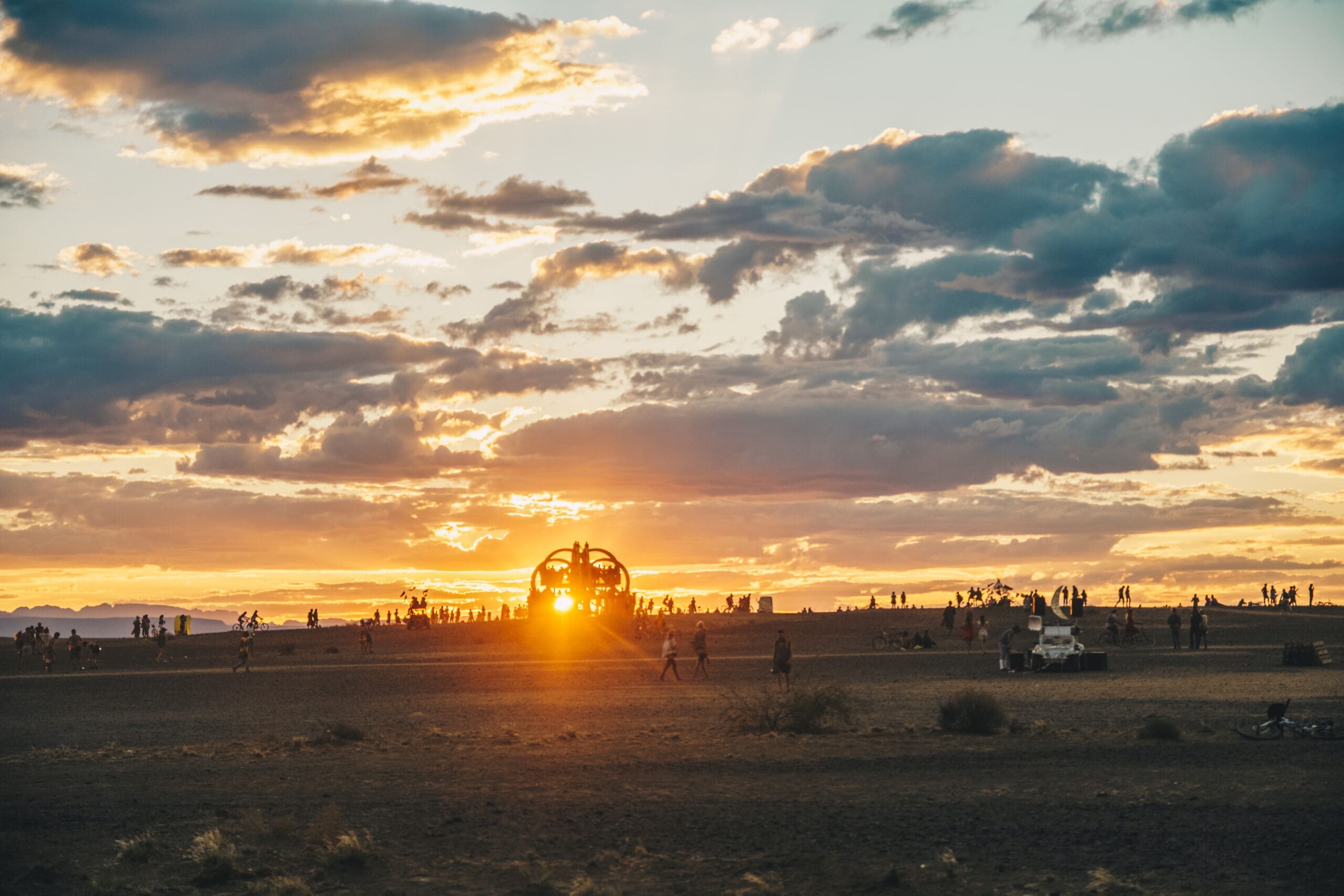 Mensen zijn verspreid over een woestijnlandschap bij zonsondergang, met dramatische wolken in de lucht. De zon schijnt achter een groot beeld in het midden, waardoor een opvallend silhouet ontstaat.