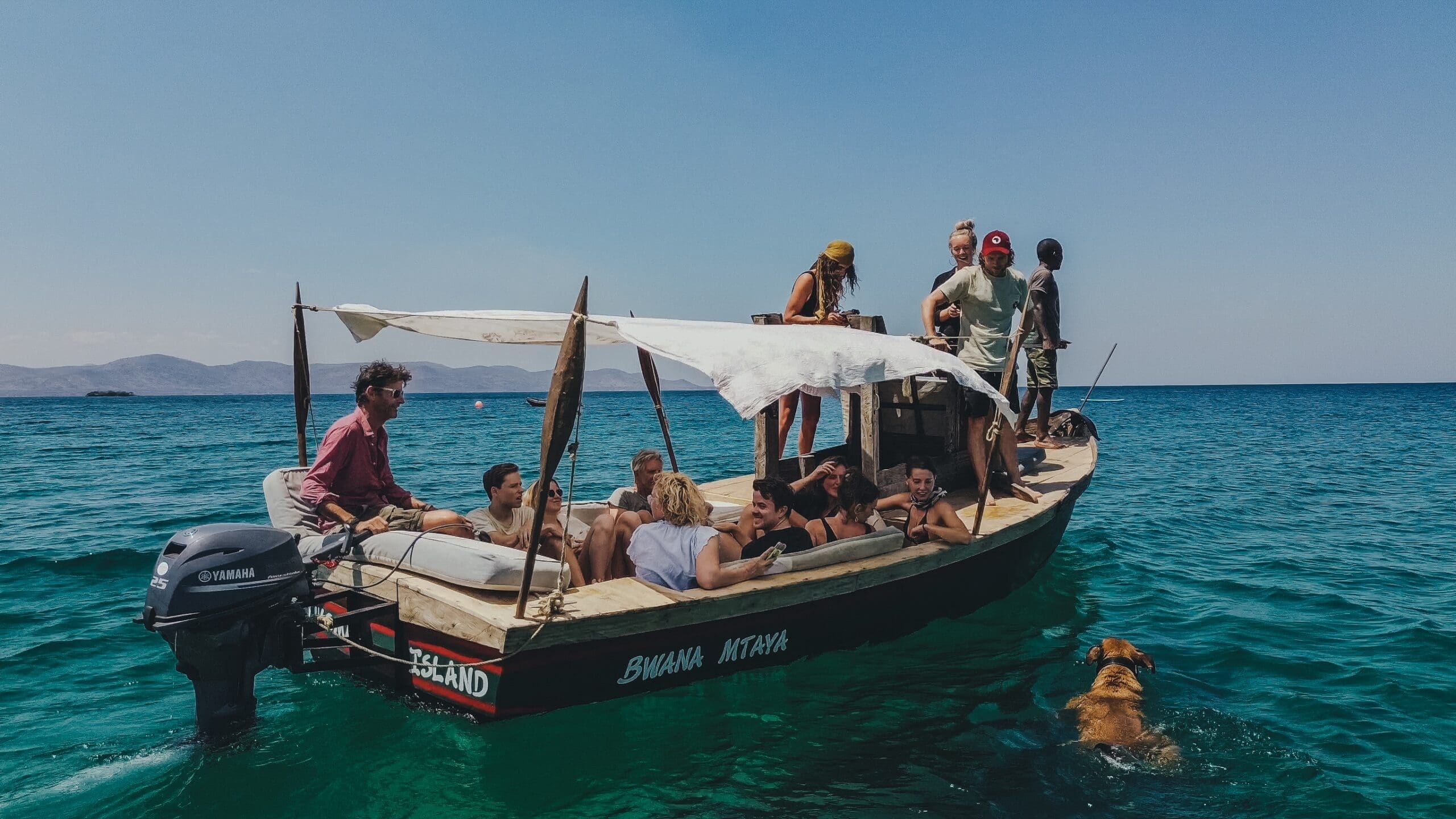 Een groep mensen ontspant zich op een houten boot met een geïmproviseerde overkapping die drijft op helderblauw water; een hond zwemt vlakbij. De lucht is helder en in de verte zijn bergen zichtbaar.