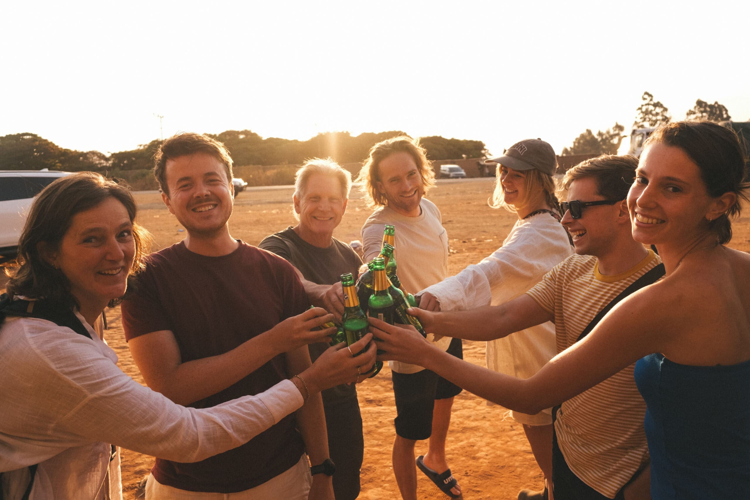 Een groep van zeven mensen die samen lachen en flessen klinken bij zonsondergang, met bomen en auto's op de achtergrond.
