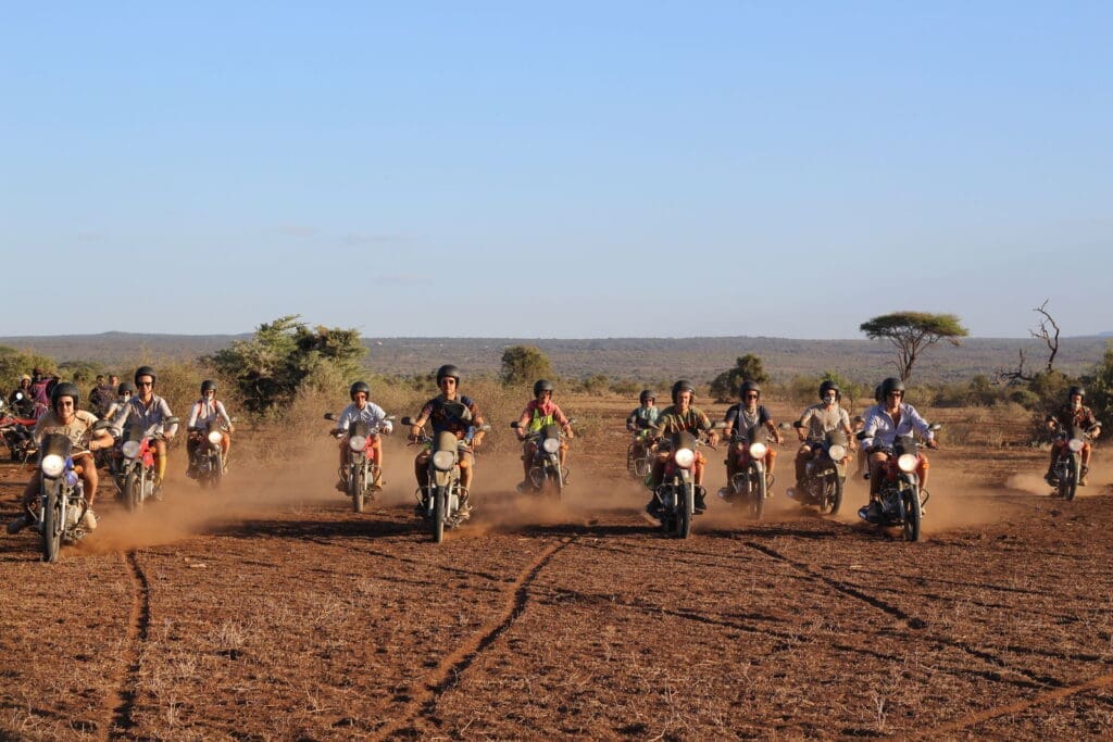 Een groep mensen rijdt op een motor door een stoffig, droog landschap met weinig bomen en een blauwe lucht op de achtergrond. De motorrijders staan verspreid in een rij, en doen stof opwaaien terwijl ze vooruit rijden.