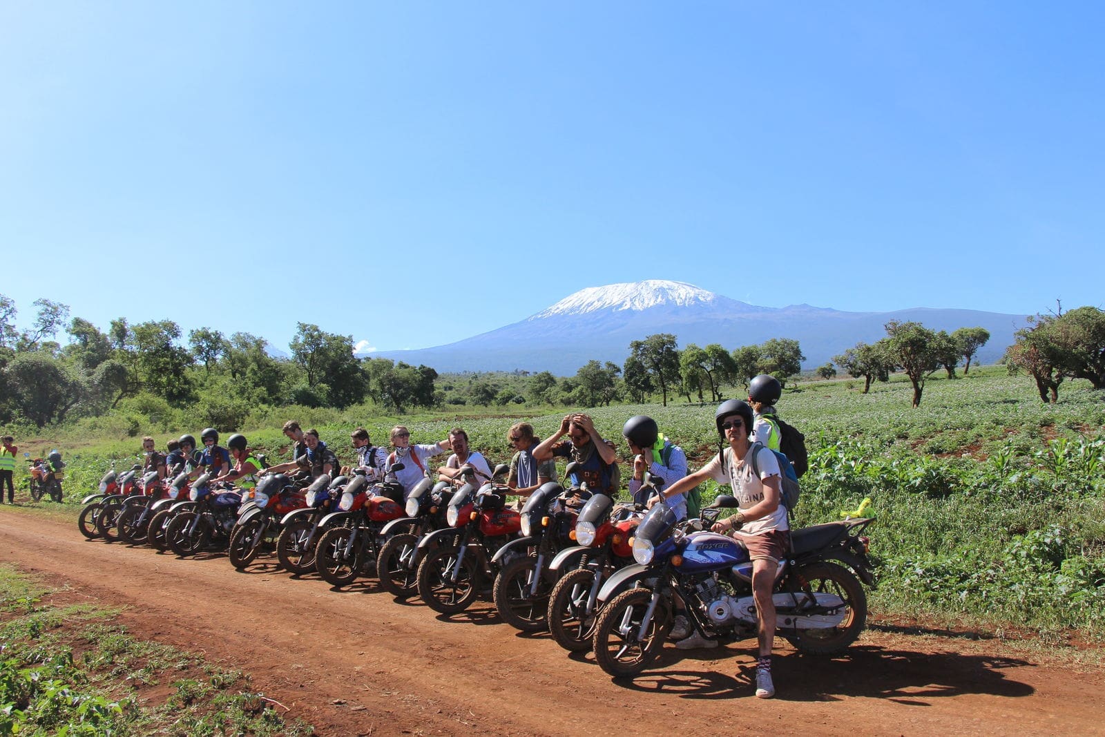 Een groep mensen met helmen op zit op motoren op een rij op een onverharde weg, met groene velden en bomen om hen heen. Een besneeuwde berg rijst op in de achtergrond onder een helderblauwe hemel.