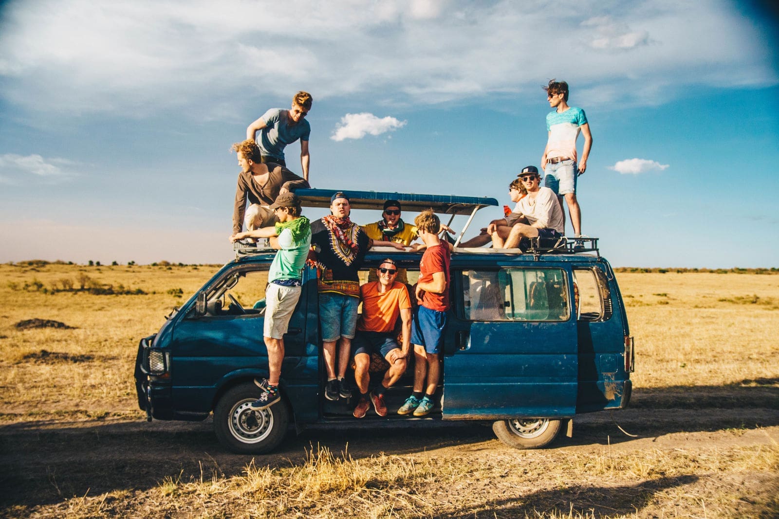 Een groep mensen poseert op en rond een blauw busje dat geparkeerd staat in een open veld onder een heldere hemel. Ze glimlachen en genieten van een zonnige dag in de buitenlucht.