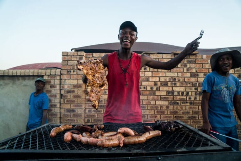 Een lachende man in een rode tanktop houdt een groot stuk gegrild vlees omhoog met een vork, staand bij een BBQ-grill met worstjes en vlees. Twee andere mensen staan vlakbij, allemaal buiten voor een bakstenen muur.