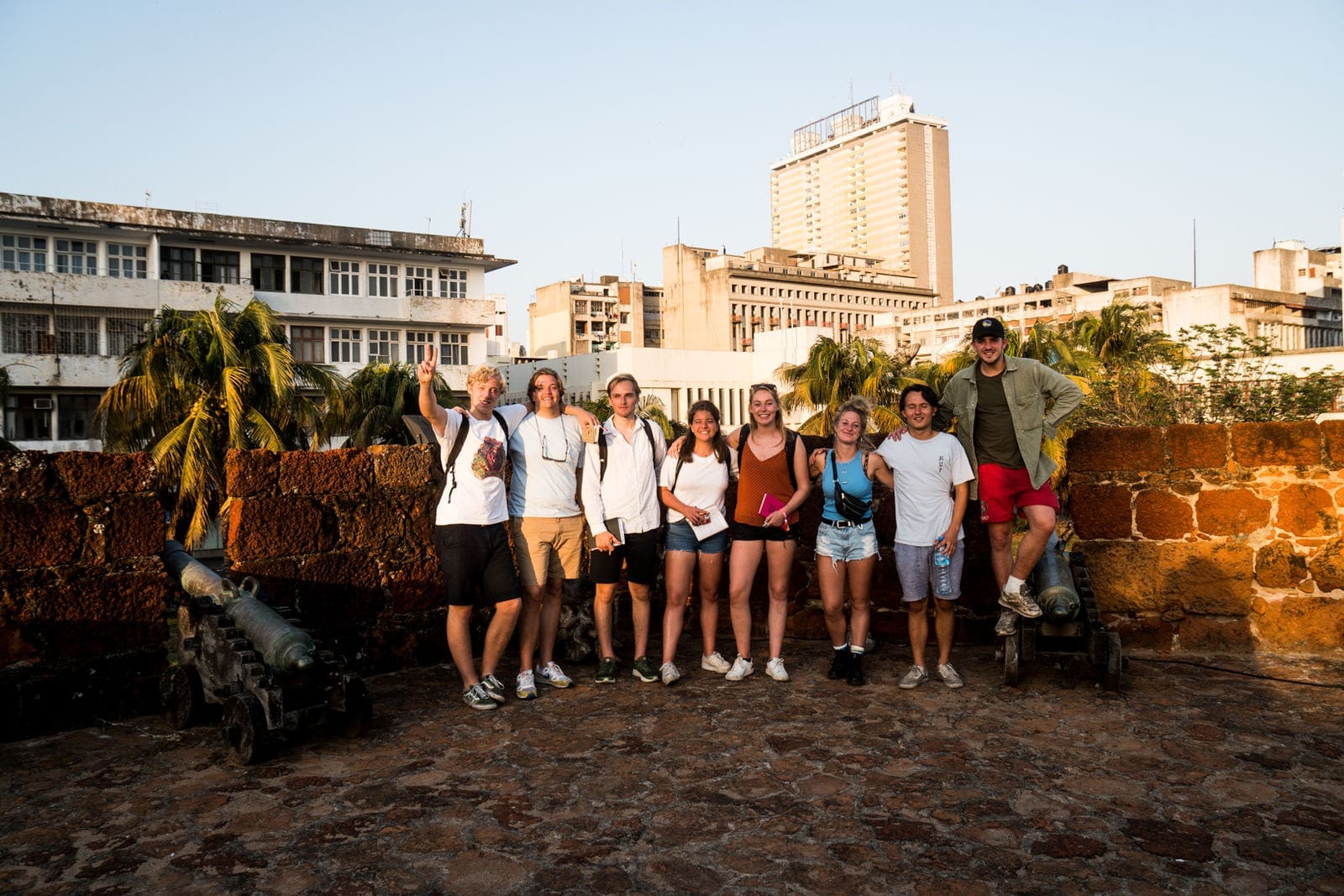 Een groep van negen mensen staat samen op een stenen vestingmuur, lachend naar de camera, met oude kanonnen aan weerszijden en hoge stadsgebouwen en palmbomen op de achtergrond.