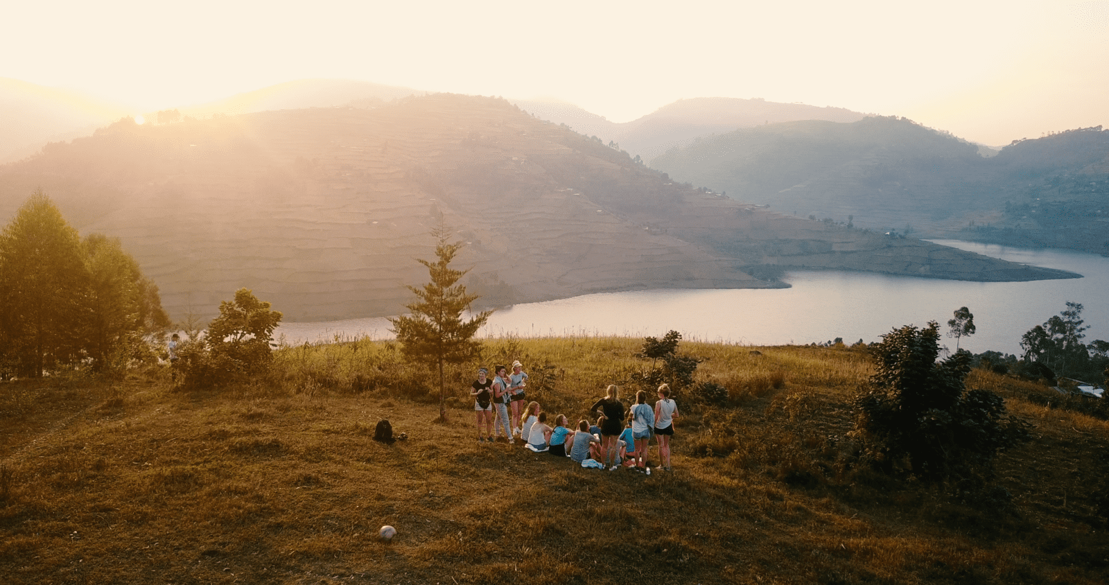 Een groep mensen verzamelt zich op een grashelling bij een meer tijdens zonsondergang, met terrasvormige bergen en zacht zonlicht op de achtergrond.