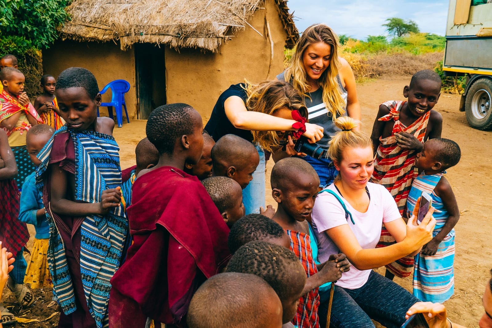 Twee vrouwen communiceren met een groep kinderen in kleurrijke traditionele kleding buiten een rieten hut. De ene vrouw zit terwijl de kinderen haar haar aanraken en de andere staat te glimlachen en te helpen. Het tafereel is levendig en vrolijk.