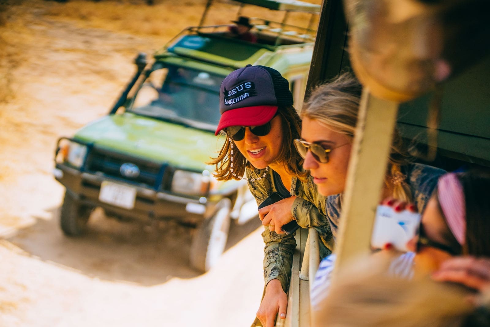 Twee vrouwen leunen uit een safarivoertuig, glimlachen en kijken naar het landschap. Op de achtergrond is een groene safari-jeep te zien op een onverharde weg. Beide vrouwen dragen een zonnebril en vrijetijdskleding.