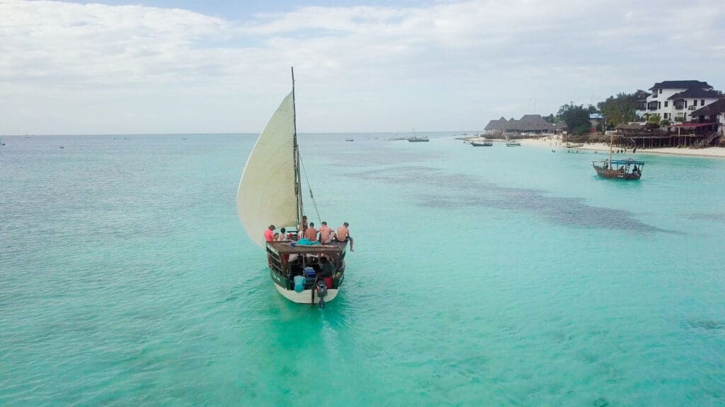 Een houten zeilboot met meerdere mensen aan boord vaart op helder turquoise water vlakbij de kust, met andere boten en gebouwen zichtbaar in de verte onder een bewolkte hemel.