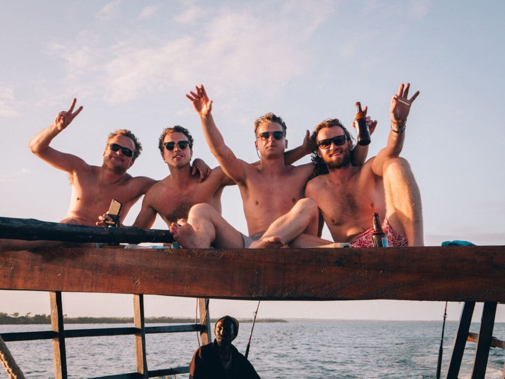 Vier mannen zitten zonder shirt op een houten constructie aan het water, glimlachen en maken vredestekens, met een drankje in hun hand. De zon gaat onder en werpt een warme gloed, en een andere persoon is gedeeltelijk zichtbaar onder hen.