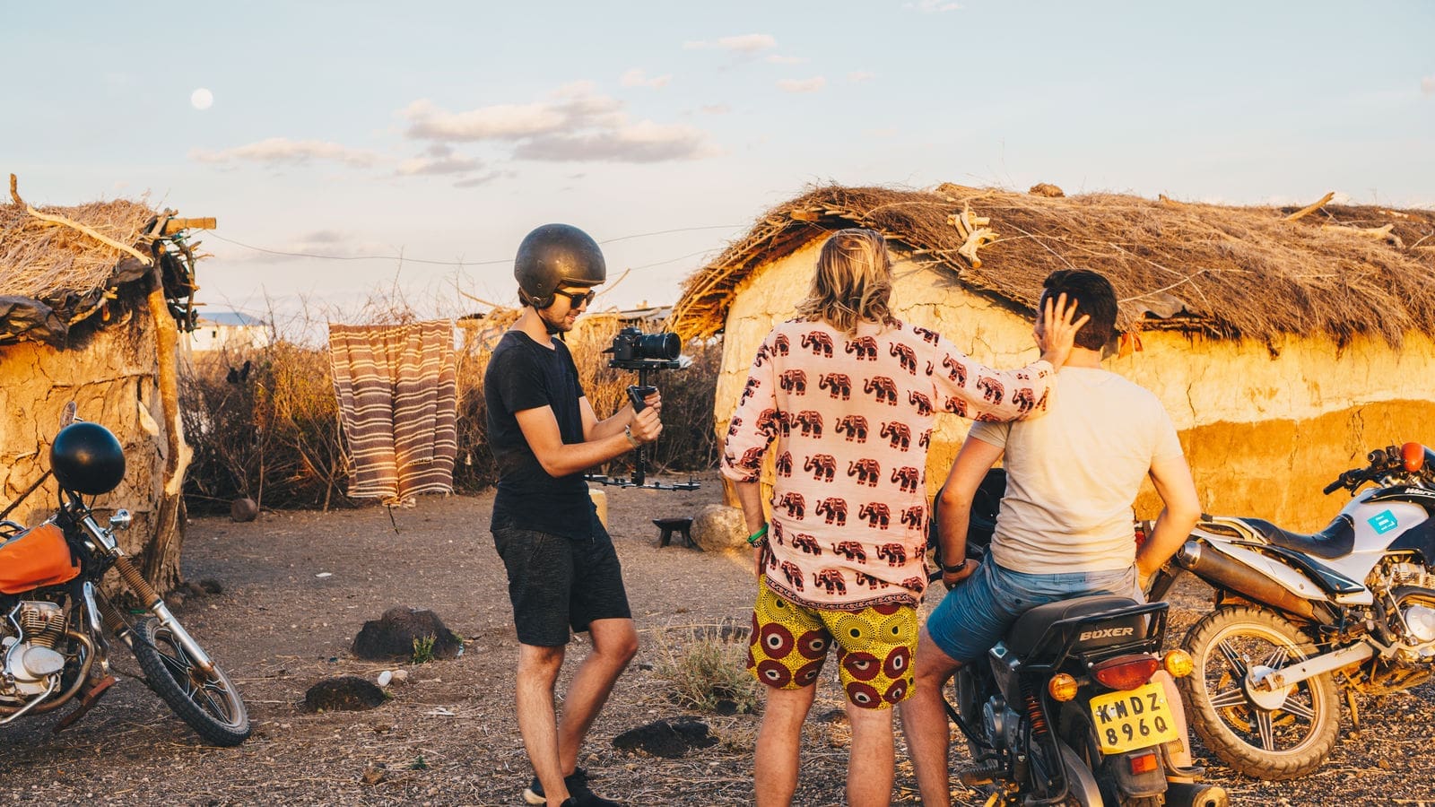 Drie mensen staan bij zonsondergang bij traditionele hutten met rieten daken; één houdt een camera vast en draagt een helm, terwijl de andere twee op een motor zitten, waarbij één persoon een hand op het hoofd van de ander laat rusten. Twee motorfietsen zijn zichtbaar.