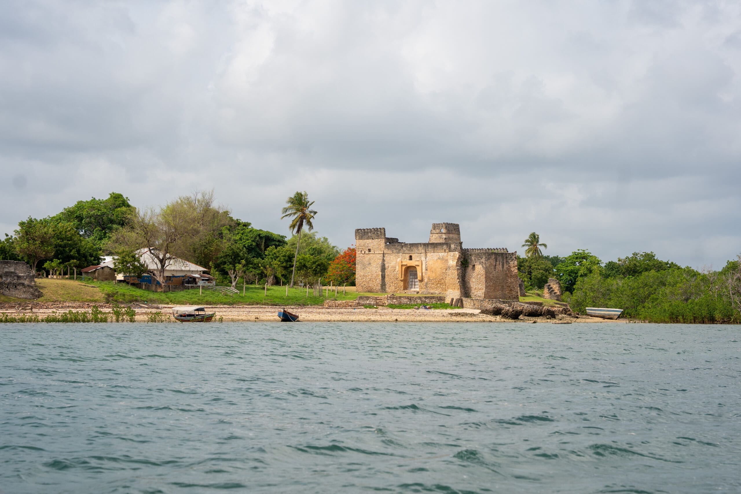 Een historisch stenen fort staat vlakbij de kustlijn, omringd door weelderig groen en een paar kleine bootjes op het water; verspreid staande bomen en een huis zijn zichtbaar in de buurt onder een bewolkte hemel.