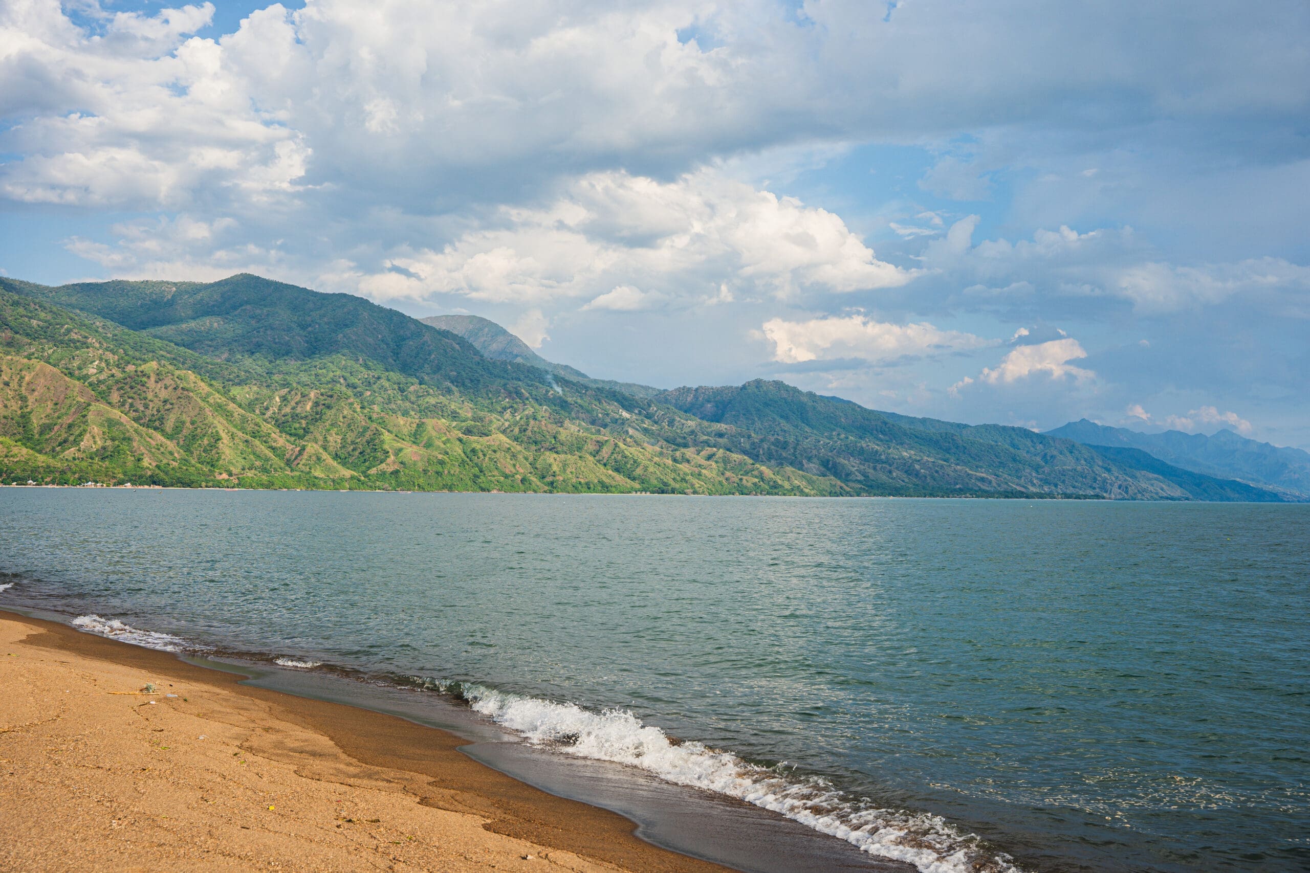 Zandstrand met zachte golven, een uitgestrekt wateroppervlak en weelderig groene bergen onder een halfbewolkte hemel.