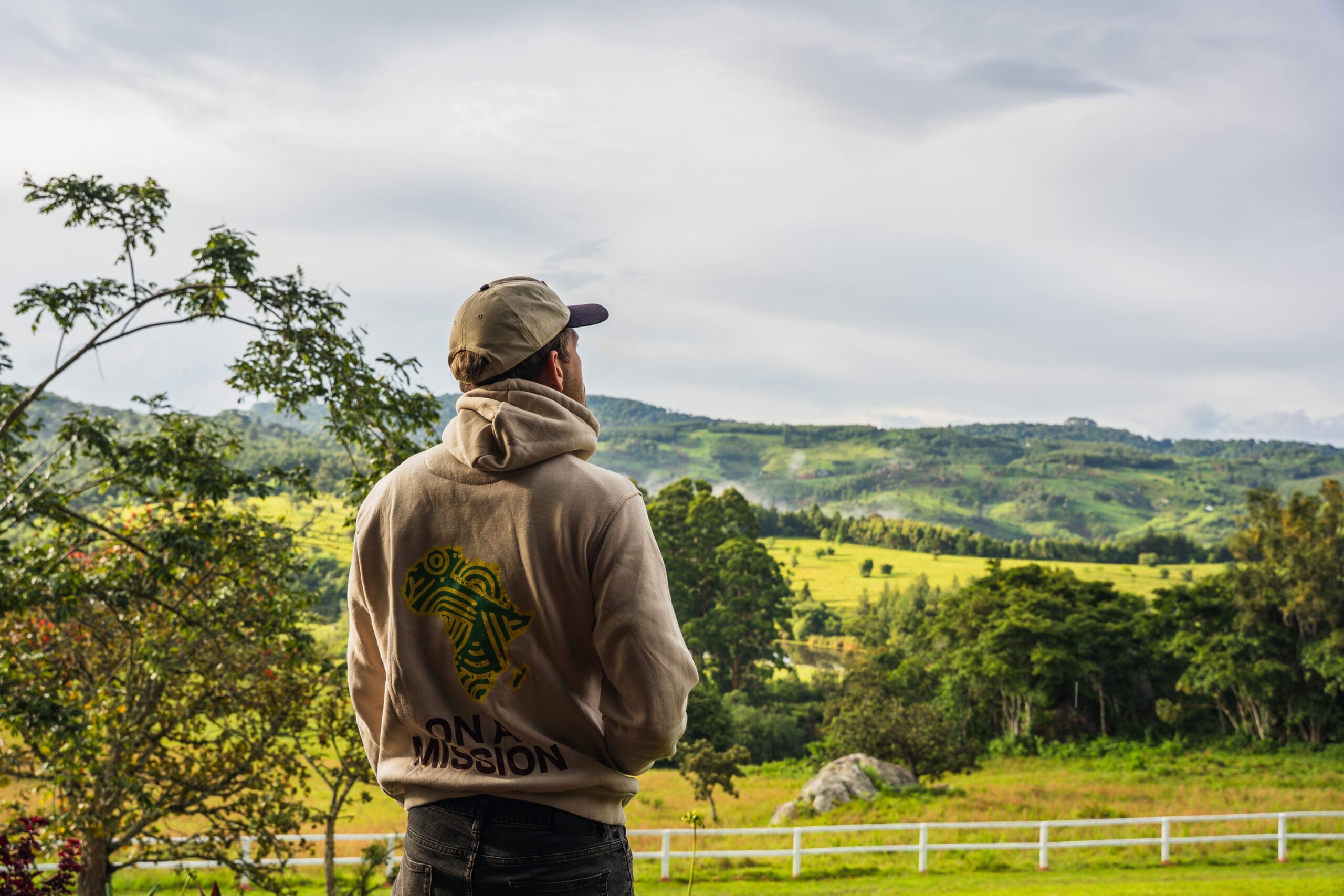 Een persoon met een beige capuchon en pet staat buiten en kijkt uit op een schilderachtig landschap van groene heuvels, bomen en een bewolkte lucht, met een wit hek zichtbaar op de voorgrond.