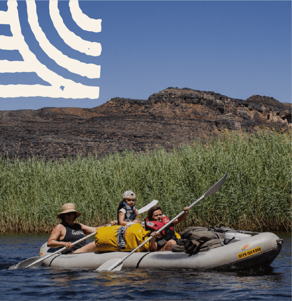 Drie mensen in zwemvesten peddelen in een geel met grijze opblaasbare kajak op een rivier, met riet langs de oever en rotsachtige heuvels op de achtergrond. Een vet wit grafisch patroon verschijnt in de lucht boven hen.