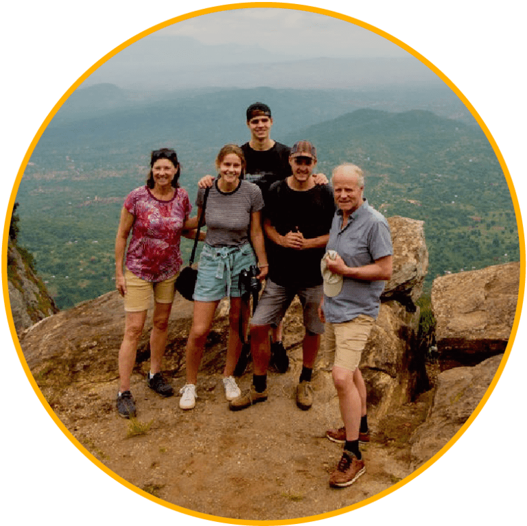 Vijf mensen staan samen op een rotsachtige richel met uitzicht op een schilderachtig, groen landschap met bergen in de verte en glimlachen naar de camera. Het lijkt een groepswandeling of sightseeing trip.
