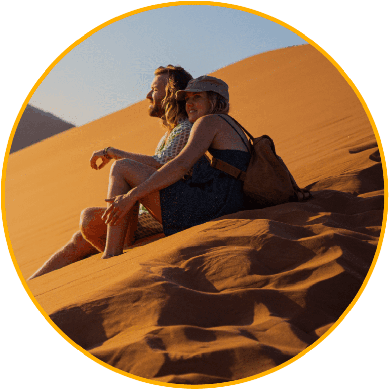 Twee mensen zitten dicht bij elkaar op het zand van een zonovergoten woestijnduin en werpen lange schaduwen. De een draagt een hoed en rugzak, terwijl de ander lang haar heeft. De sfeer is ontspannen en warm.