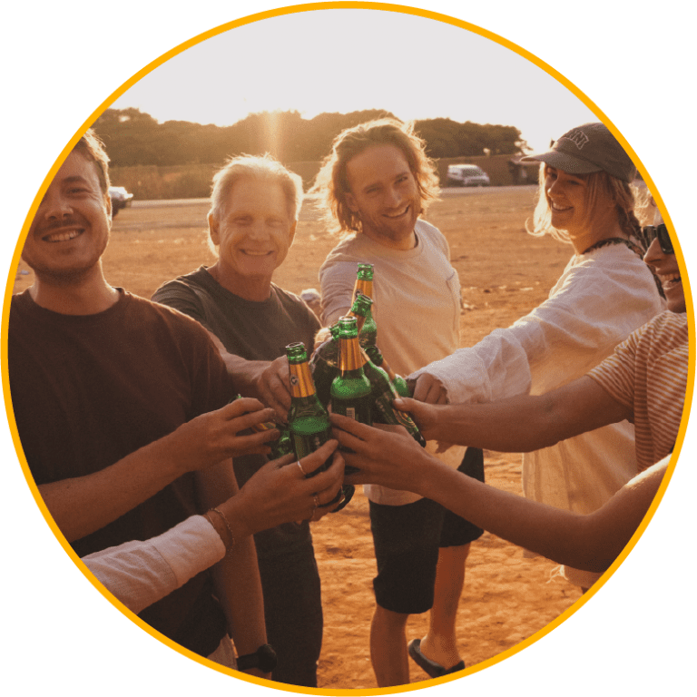 Een groep mensen die glimlachen en samen bierflesjes klinken bij zonsondergang, genietend van een informeel samenzijn of feest.