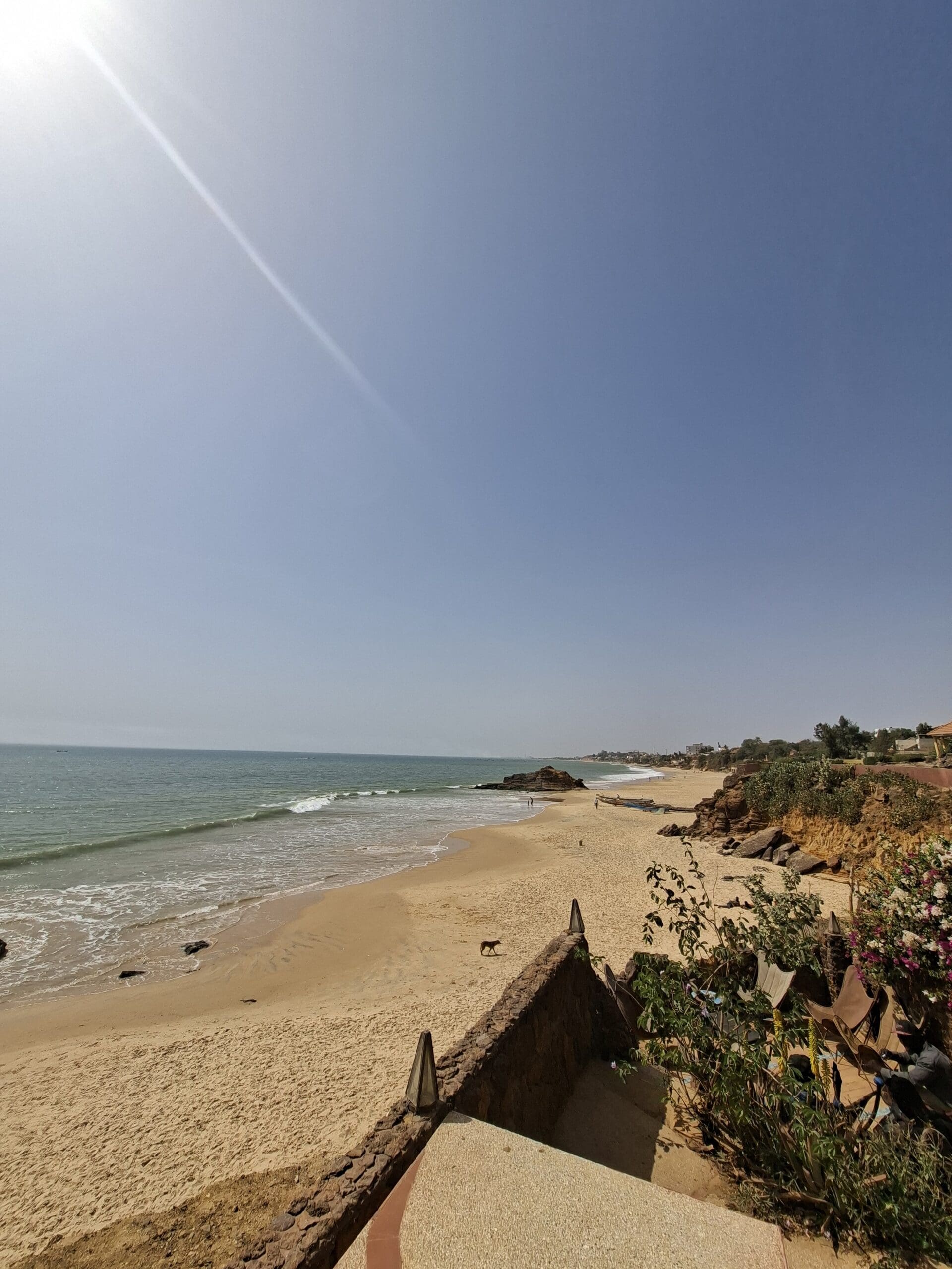 Een zonnige, heldere dag op een zandstrand met zachte golven, rotsen en een paar planten op de voorgrond. De kustlijn kromt zich in de verte onder een blauwe hemel met een zonnestraal zichtbaar.