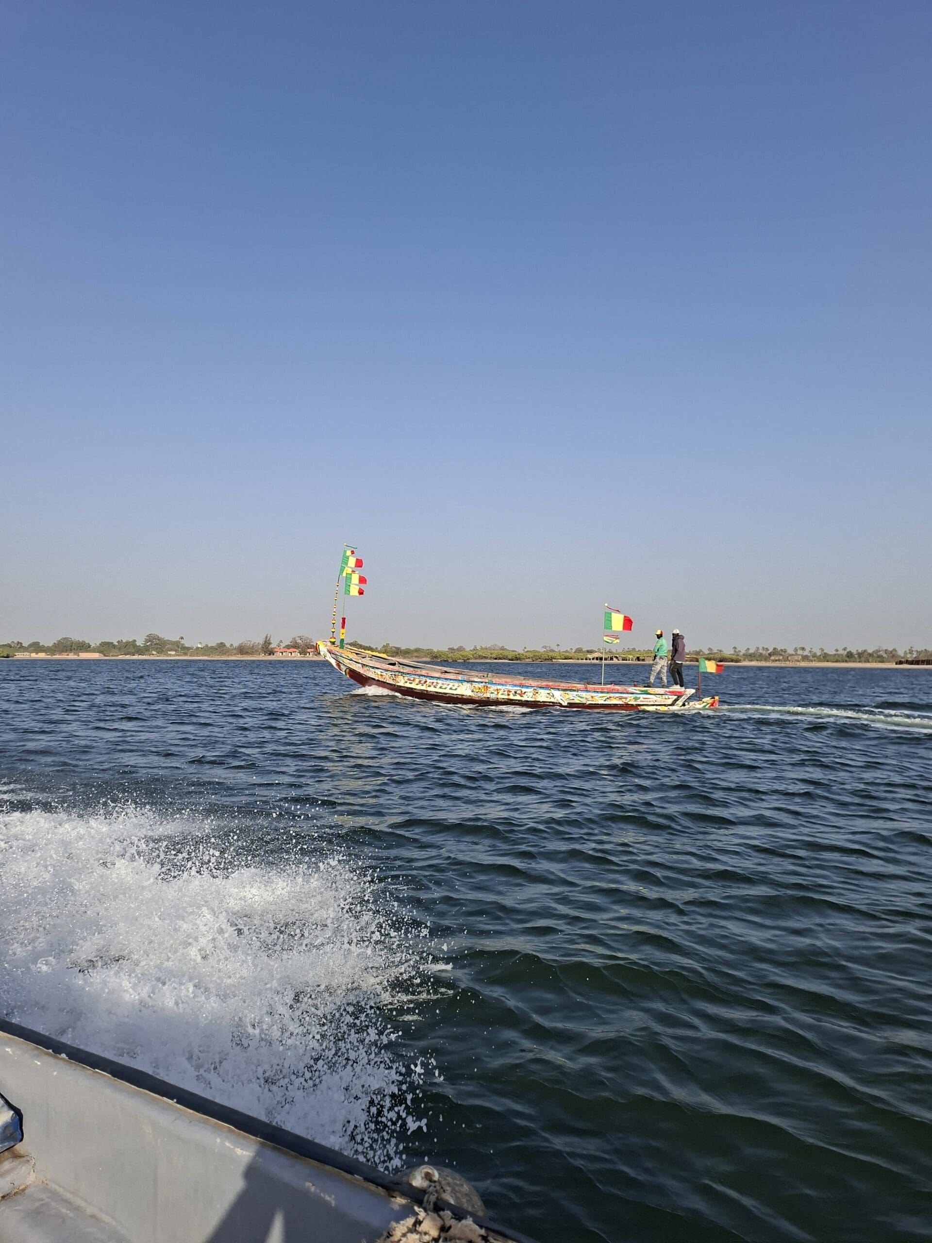 Een kleurrijke houten boot met vlaggetjes drijft op een kalme rivier onder een strakblauwe hemel. Een persoon staat op de boot, terwijl water opspat van een andere boot op de voorgrond.