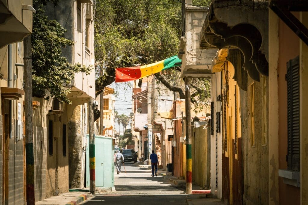 A narrow street lined with old buildings, colorful green, yellow, and red flag hanging overhead, with a few people walking in the distance under dappled sunlight.