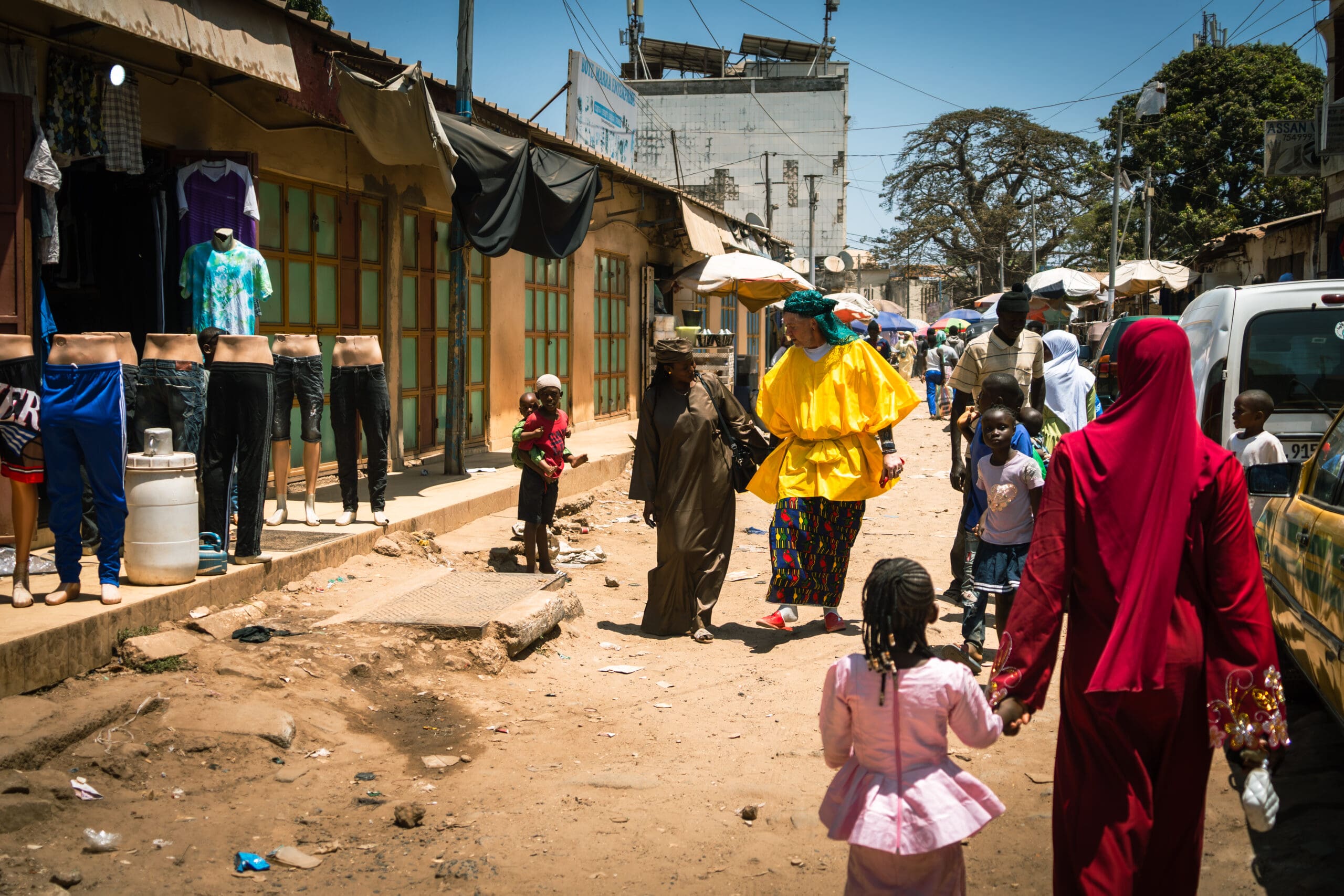 Een drukke marktstraat met lopende mensen, sommigen met felgekleurde kleding. Winkels tonen broeken op paspoppen buiten. Het tafereel is levendig, met kinderen en volwassenen onder een zonnige, heldere hemel.