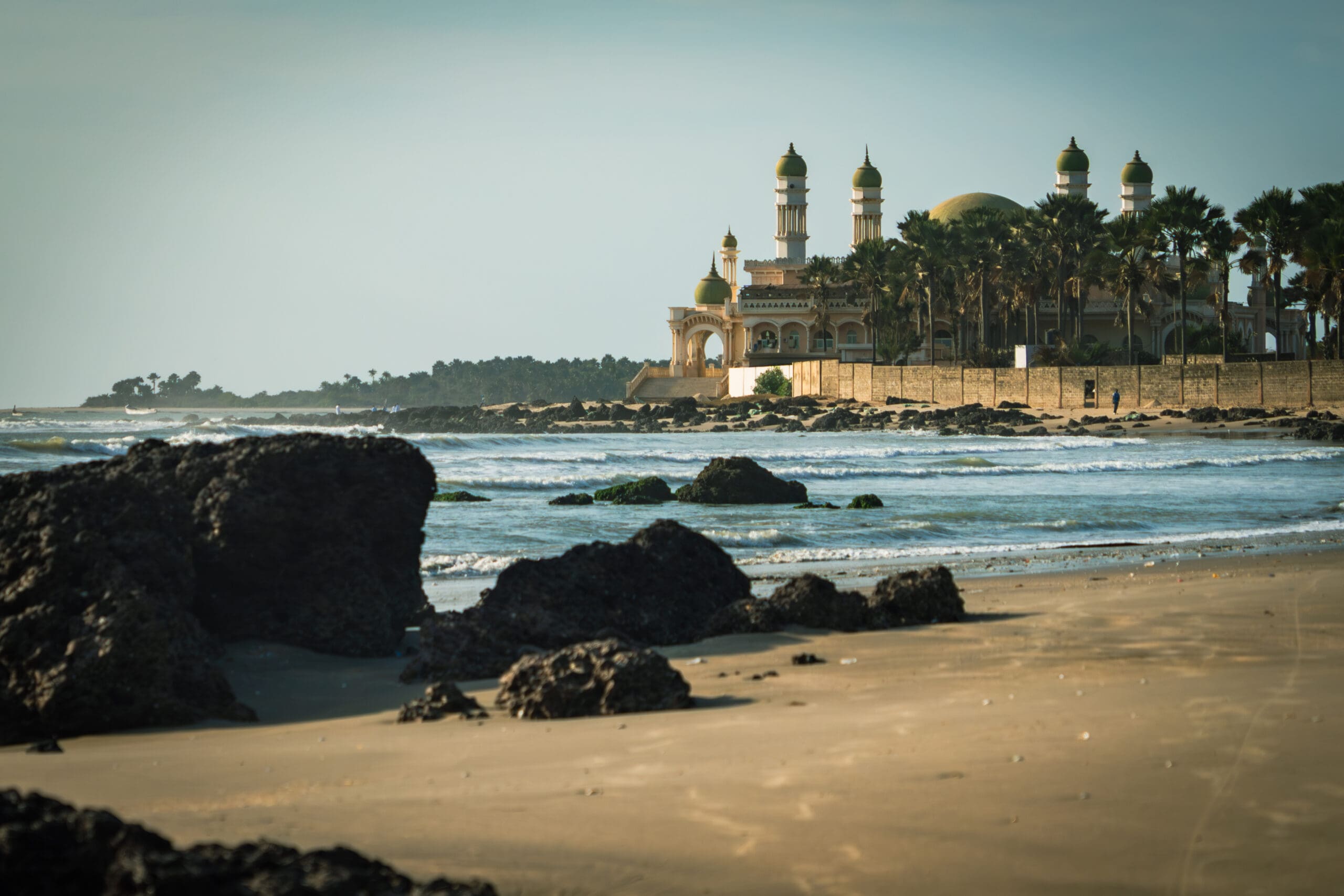 Gouden koepelmoskee met meerdere torens omringd door palmbomen, gelegen aan de kust met golven en donkere rotsen verspreid langs het zandstrand op de voorgrond. De lucht is helder.