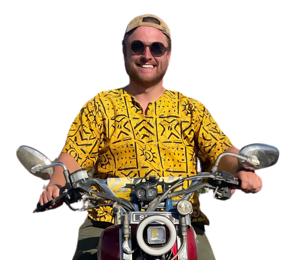 Een lachende man met zonnebril, beige pet en felgeel overhemd met patroon zit op een motorfiets, houdt het stuur vast, met een witte achtergrond.