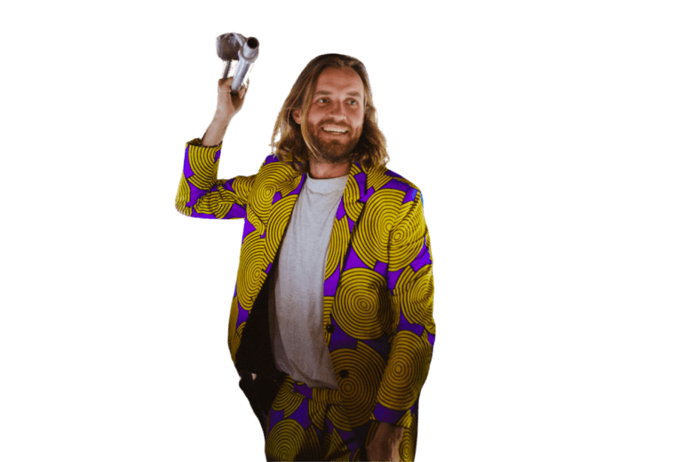Een lachende man met lang haar draagt een felgeel en paars gedessineerd pak en houdt een zilveren camera boven zijn schouder. De achtergrond is egaal wit.