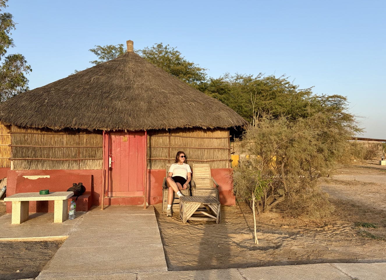 Een persoon zit op een houten stoel buiten een rietgedekte hut met rode deur, omgeven door zandgrond, bomen en een stenen bank onder een helderblauwe hemel bij zonsondergang.