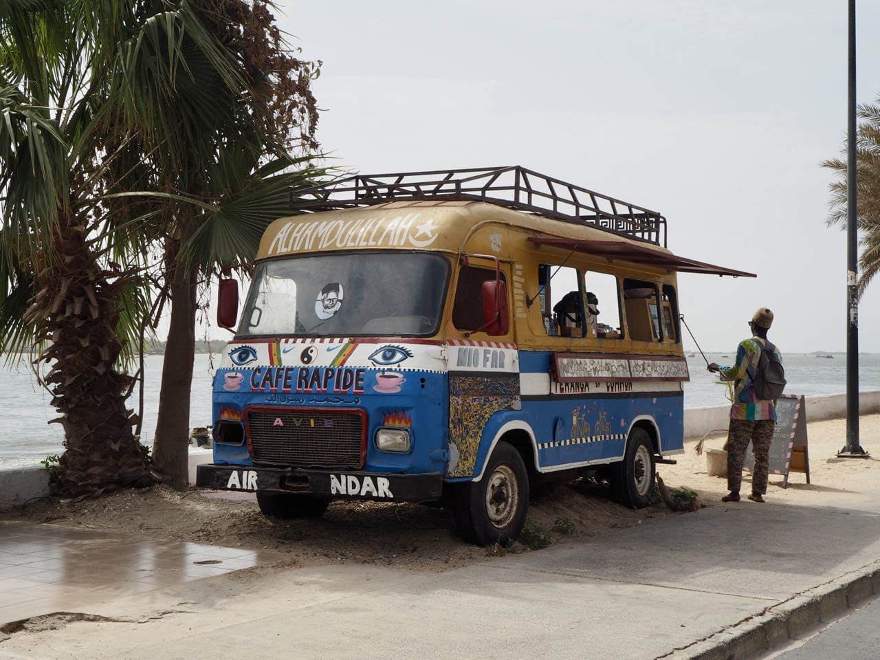 Een kleurrijke, vintage voedselwagen met de naam Café Rapide staat geparkeerd aan het strand onder palmbomen. Een persoon staat vlakbij op de stoep en kijkt in de richting van de zee. De truck is beschilderd met felle patronen en tekst.