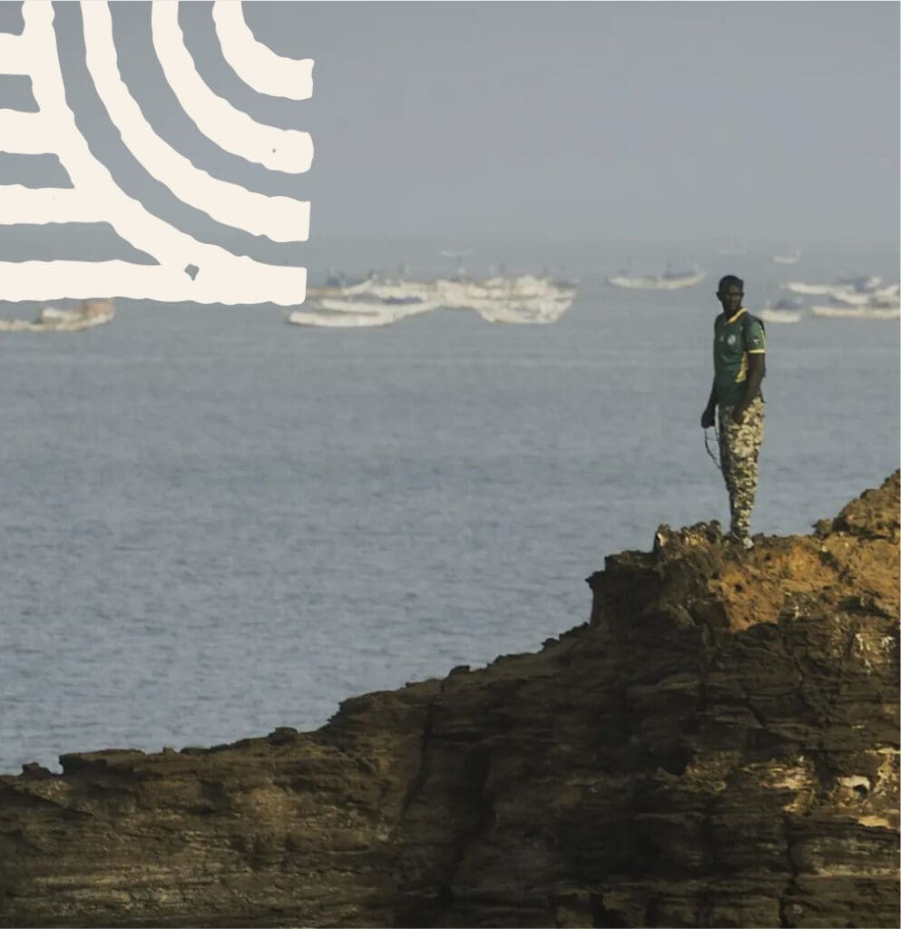 Een persoon in camouflagebroek staat op een rotsachtige klif met uitzicht op de oceaan, met meerdere boten zichtbaar op het water en een wit abstract patroon in de lucht.