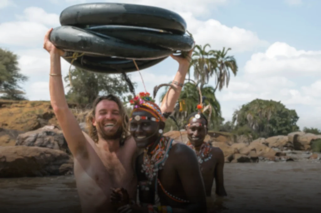 Een lachende man zonder shirt houdt rubberen binnenbanden boven zijn hoofd terwijl hij in een rivier staat met twee mensen in kleurrijke traditionele kleding, met bomen en rotsen op de achtergrond.