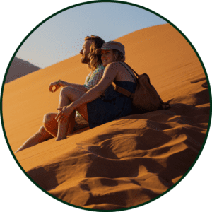 Zwei Menschen sitzen dicht beieinander auf einer sonnenbeschienenen Sanddüne, lächelnd und entspannt. Der eine trägt einen Hut und einen Rucksack, der andere hat lange Haare. Der Himmel ist klar, und ihre Schatten fallen auf den strukturierten Sand.