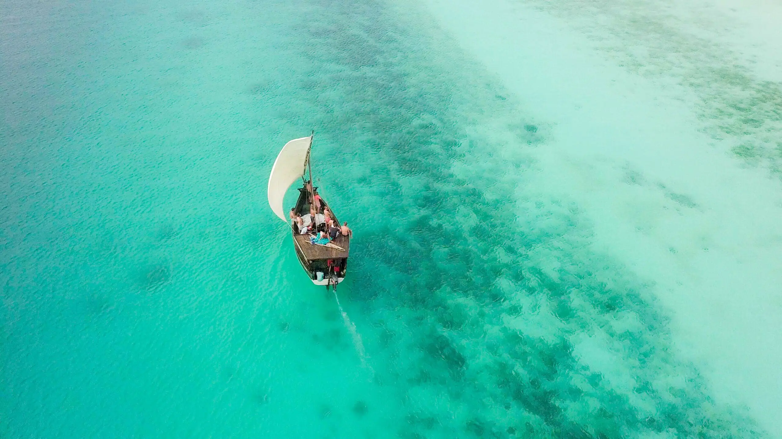 A small sailboat with several people aboard glides through clear turquoise water, leaving a gentle wake behind, surrounded by shallow patches and deeper blue areas in the sea.
