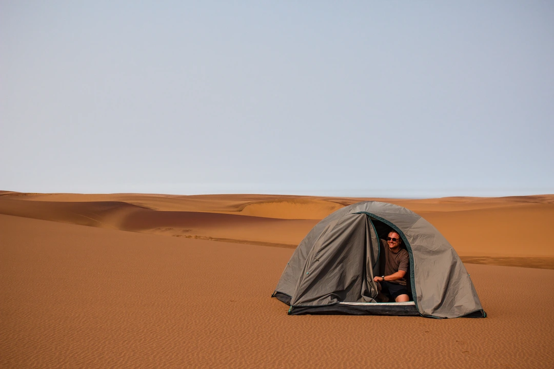 Een persoon zit in een grijze tent die is opgezet op het oranje zand van een woestijn, omringd door zandduinen onder een heldere hemel.