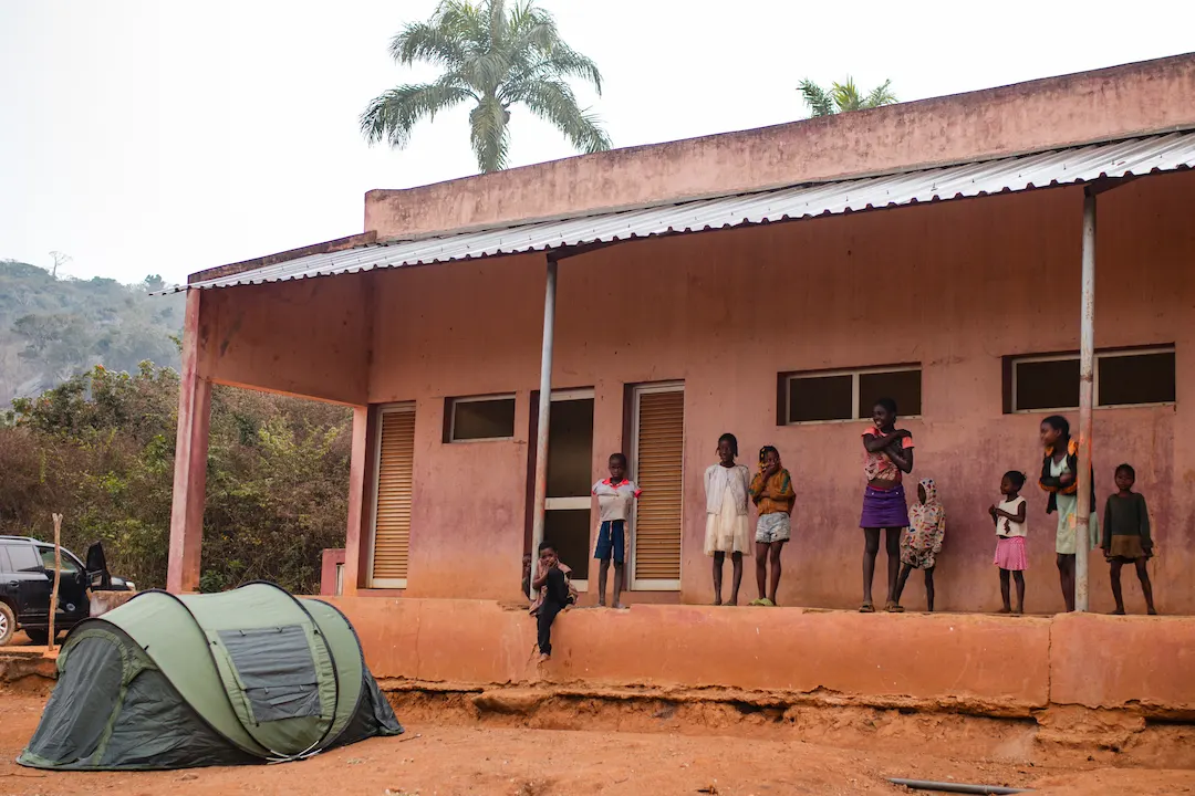 Een groep kinderen staat op de veranda van een verweerd gebouw met een metalen dak. Een groene tent staat op de zandgrond ervoor en een zwarte auto is gedeeltelijk zichtbaar aan de linkerkant. Palmbomen en heuvels zijn op de achtergrond te zien.
