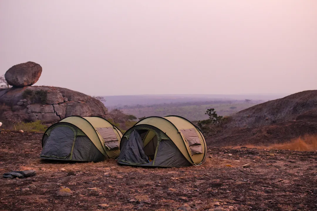 Twee groene tenten opgezet op een rotsachtige ondergrond bij zonsondergang met grote rotsblokken en verre wazige heuvels op de achtergrond, waardoor een vredige kampeerscène in de buitenlucht ontstaat.