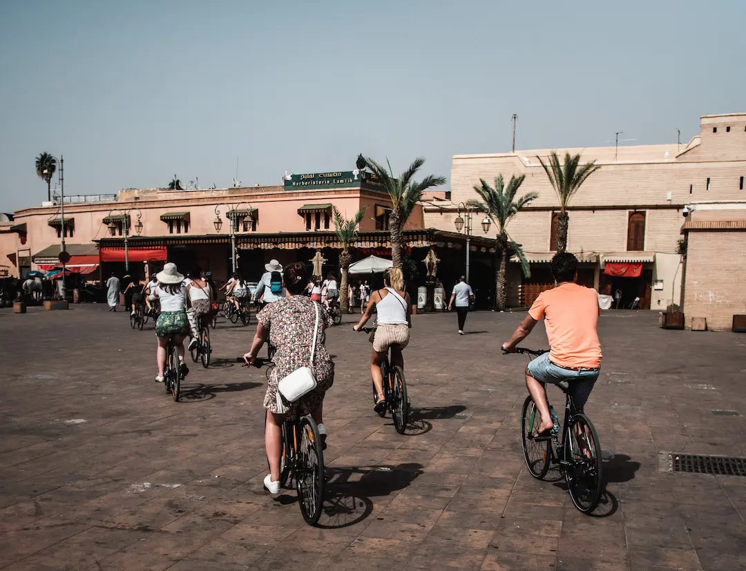 Een groep mensen fietst over een zonnig, open plein met palmbomen en beige gebouwen op de achtergrond. Sommige fietsers dragen hoeden en zomerkleding.