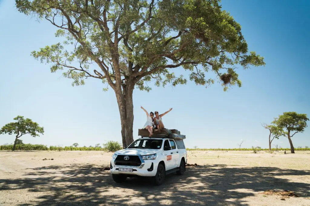 Two people sit on the roof of a white SUV parked under a large tree in a sunny, open landscape, raising their arms in celebration. The sky is clear and blue, and the area appears remote and natural.